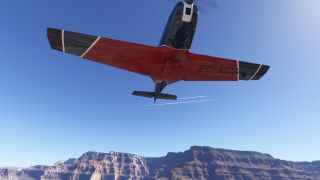 A red and black propeller aircraft passes overhead, showing its underside above rocky canyon cliffs beneath a clear blue sky.