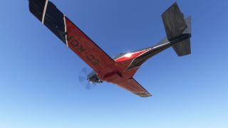 A red and black single-engine aircraft flies overhead against a clear blue sky, showing its underside and spinning propeller.