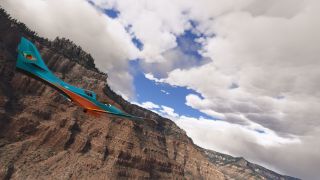 A turquoise and orange aircraft banks alongside steep rocky cliffs, with scattered trees and large clouds filling the sky.