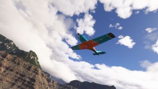 A turquoise and orange single-engine aircraft flies overhead above rocky, forested cliffs beneath a blue sky filled with white clouds.