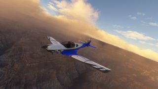 A blue and white single-engine aircraft with checkered markings banks over rugged canyon terrain beneath golden clouds.