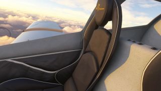 A black cockpit seat with white piping sits beside a canopy window overlooking a white wing and sunlit clouds.