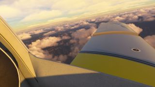 A cockpit side window frames a yellow-and-gray aircraft wing above scattered clouds and rugged terrain in warm sunlight.