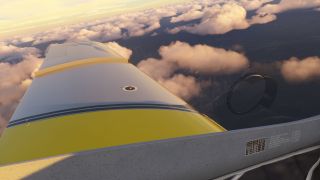 A cockpit side window overlooks a yellow-and-white wing with dark stripes above scattered clouds illuminated by warm sunlight.
