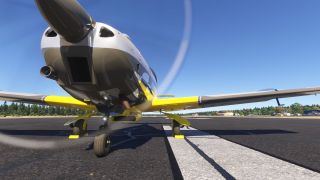 A low frontal view shows a yellow, gray, and white propeller aircraft with extended landing gear and yellow wheel chocks on marked pavement.