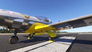 A silver, black, and yellow aircraft sits on runway markings with wheel chocks fitted and a pilot visible in the cockpit.