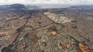 Dense urban neighborhoods, intersecting highways, and white-roofed industrial buildings extend toward a coastal bay beneath a flat-topped mountain.