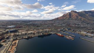 Cargo ships line harbor quays beside container yards, while a dense city stretches toward a flat-topped mountain beneath scattered clouds.