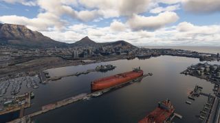 A large red tanker sits moored in a harbor surrounded by marinas, storage tanks, and a city beneath flat-topped mountains.