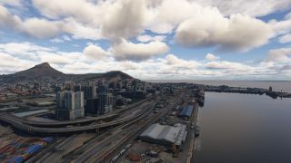 A coastal city stretches beside a harbor, with elevated highways, waterfront warehouses, high-rise buildings, and mountain slopes beneath scattered clouds.