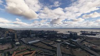 A coastal city stretches around railway yards and elevated highways, with cargo ships and cranes lining the harbor beneath scattered clouds.