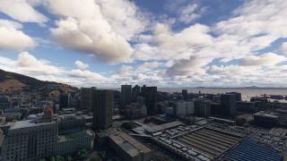 High-rise buildings overlook a rail terminal and harbor, with hillside neighborhoods to the left beneath scattered clouds.