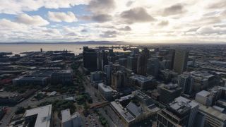 A coastal city skyline overlooks harbor cranes and docks, with tree-lined streets below and distant hills beneath scattered clouds.