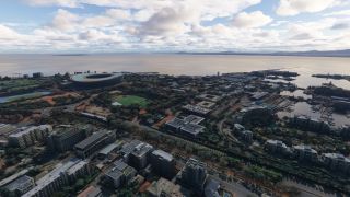 An aerial view shows a coastal city with an oval stadium, tree-lined roads, and harbor basins beneath scattered clouds.