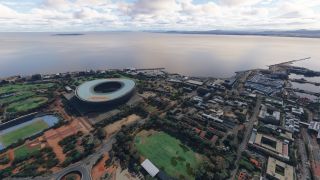 A large oval stadium stands beside sports fields and coastal city streets, with harbor docks and a broad bay beyond.