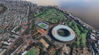 A large oval stadium stands beside a blue athletics track, golf course, and dense coastal neighborhoods along the waterfront.
