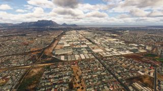 An aerial view shows dense residential blocks and large industrial warehouses, with a flat-topped mountain and coastline beneath scattered clouds.
