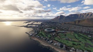 An aerial view shows a coastal city with an oval stadium, harbor basins, waterfront buildings, and a flat-topped mountain beneath scattered clouds.