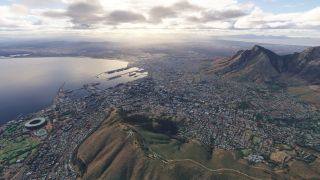 A coastal city spreads around a harbor and oval stadium, with steep mountains rising beside dense neighborhoods under scattered clouds.