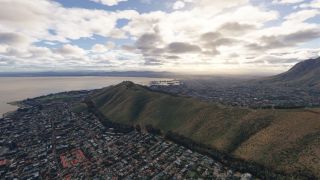 A coastal city surrounds a broad grassy ridge, with a waterfront stadium, distant harbor, and mountains beneath scattered clouds.