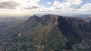 A flat-topped mountain rises above dense urban neighborhoods, with steep rocky cliffs, winding hillside trails, and distant ocean beneath scattered clouds.