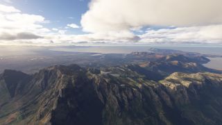 Steep mountain cliffs and hilltop reservoirs overlook a sprawling coastal city, with a broad bay and scattered clouds beyond.