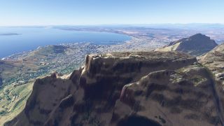 A flat-topped mountain with steep rocky cliffs overlooks a dense coastal city, harbor piers, and a broad blue bay.