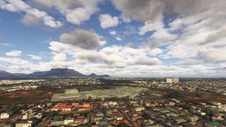 A sprawling city surrounds a large flat-roofed complex, with a broad mountain ridge rising beneath scattered clouds in the distance.