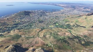 A dense coastal city spreads between grassy mountain slopes and a broad blue bay, with harbor docks and an offshore island visible.