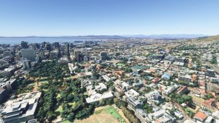 A coastal city spreads around a tree-filled park, with downtown towers, a harbor, hillside neighborhoods, and distant mountains beneath clear skies.