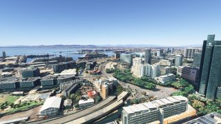 An aerial view shows waterfront high-rises, curving elevated roads, and a harbor filled with ships and cranes beneath clear blue skies.