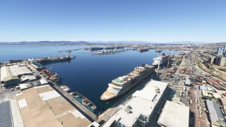 Two cruise ships dock beside warehouses and container yards, while cargo vessels and cranes line the opposite side of the harbor.