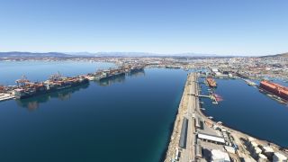 Container ships sit beside dockside cranes in a broad harbor, with industrial piers, storage tanks, and a sprawling city beyond.
