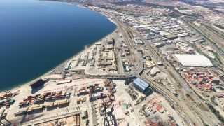 An aerial view shows a coastal industrial port with stacked shipping containers, cranes, warehouses, and railway tracks beside a blue bay.