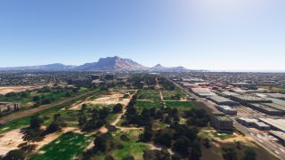An aerial view shows tree-dotted open ground and industrial buildings, with dense urban development stretching toward a broad, flat-topped mountain.