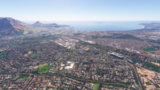 Dense urban neighborhoods spread toward a broad bay, with mountains on the left, harbor docks, and intersecting runways near the shoreline.