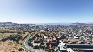 A coastal city stretches around a broad bay, with curving highways, red-roofed buildings, grassy hillsides, and harbor cranes visible.