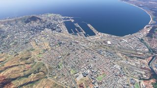 A coastal city surrounds a large harbor with long piers, rail yards, dense neighborhoods, and grassy hills beside a curved bay.