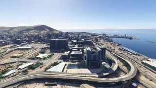 Elevated highways curve around high-rise buildings beside a harbor, with railway tracks, dockside warehouses, and hills visible under clear skies.