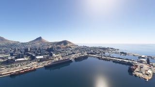 Large ships line a sheltered harbor beside dense city buildings, with dry mountain slopes rising behind the waterfront under clear skies.