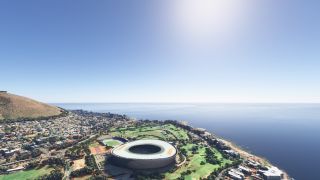 An oval stadium stands beside green parkland and dense coastal neighborhoods, with a dry hillside overlooking the ocean.