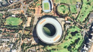 An overhead view shows an oval stadium with DHL roof logos, surrounded by sports fields, golf fairways, roads, and buildings.