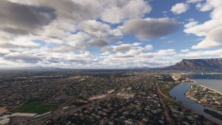 An aerial view shows dense urban neighborhoods, a distant runway, coastal waterways, and a flat-topped mountain beneath scattered clouds.
