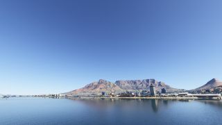 Harbor cranes and waterfront buildings line calm blue water beneath a broad, flat-topped mountain and clear sky.