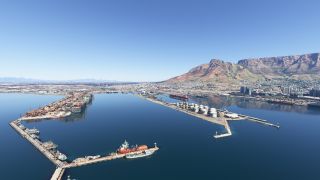 A harbor contains docked ships, container cranes, and cylindrical storage tanks, with dense urban development beneath a broad, flat-topped mountain.