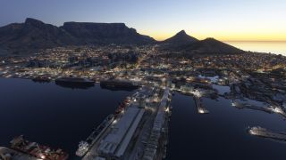 Cape Town’s illuminated harbor contains docked ships and long warehouses, with city buildings beneath Table Mountain against a golden horizon.