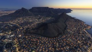 Illuminated streets spread around Cape Town’s mountain ridges, with flat-topped Table Mountain beyond and the ocean beneath a golden horizon.