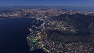 Illuminated streets spread around a coastal harbor and oval stadium, while steep mountains rise above the city at dusk.