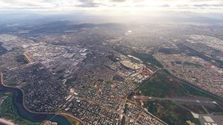 An aerial view shows dense urban neighborhoods, industrial buildings, a winding river, and an airport runway beneath hazy mountains and scattered clouds.