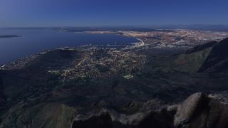 A coastal city stretches around a broad bay at dusk, with illuminated streets, harbor facilities, and steep mountain slopes in the foreground.